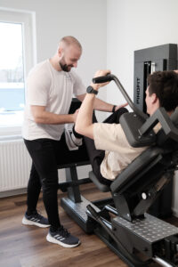 Ein Fitness-Trainer hilft einem Klienten beim Beinpressen auf einem PinoFit-Gerät im Fitnessstudio.