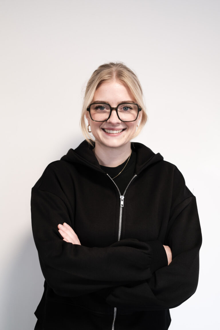 Lächelnde Frau mit Brille, schwarzem Hoodie und Kopfhörern, die mit verschränkten Armen posiert. Ideal für Mode- und Lifestyle-Themen Physiowell Westerstede.